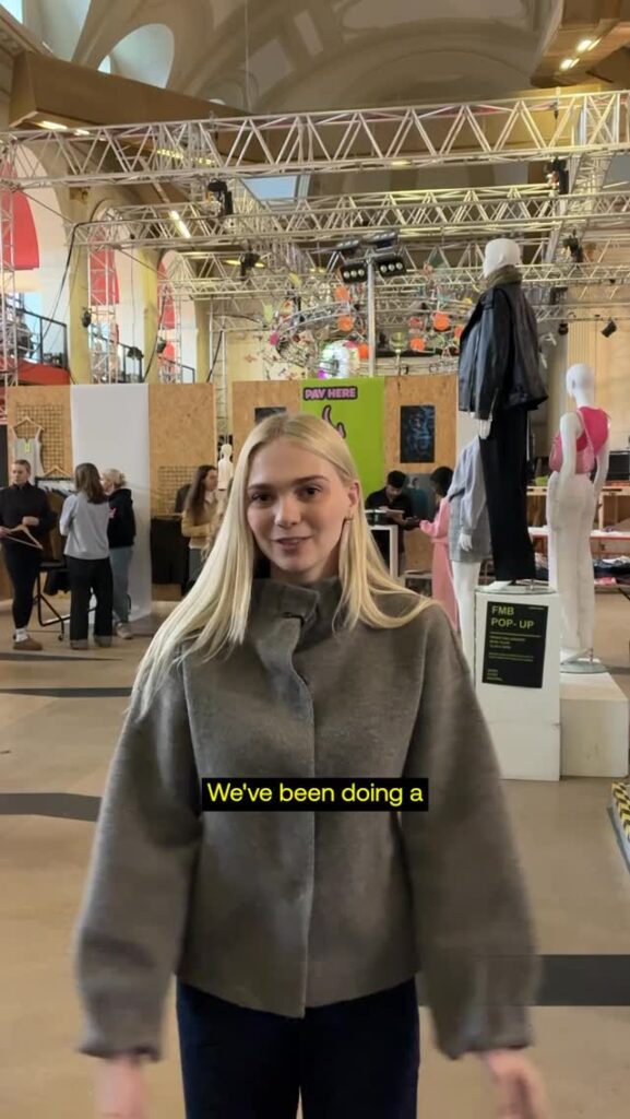 A blonde woman in a gray coat stands indoors at an event with people, mannequins, and clothing displays in the background. Yellow captions at the bottom read, "We've been doing a.