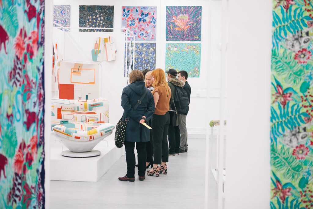 A group of people stand closely together, viewing colorful abstract sculptures in a modern art gallery. Patterned fabric panels and vibrant paintings decorate the bright, white-walled space.