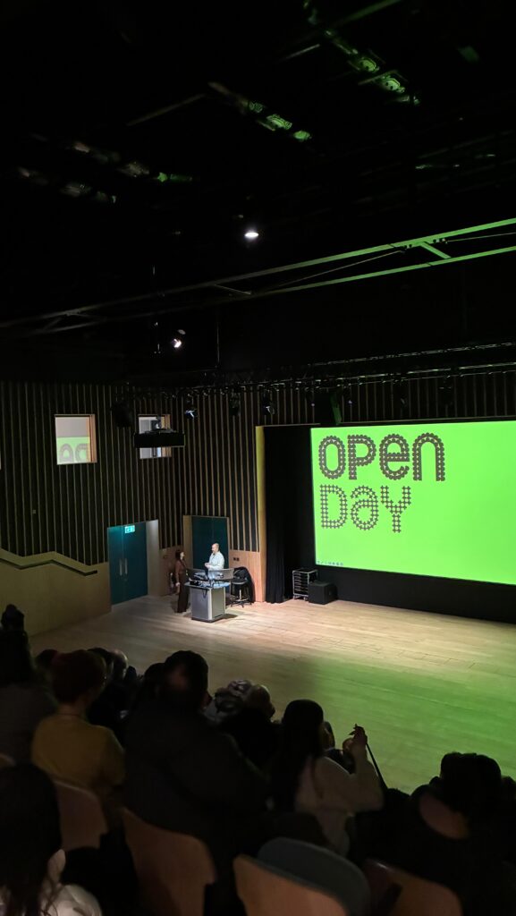 A person stands at a podium on a stage in a dimly lit auditorium, addressing an audience. A large green screen behind them displays the words "OPEN DAY" in bold letters.