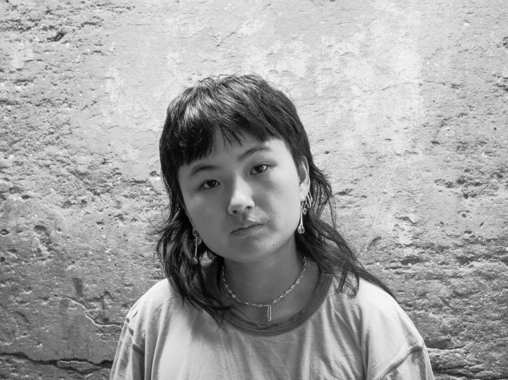 A person with medium-length dark hair and bangs, wearing a T-shirt, earrings, and a necklace, looks at the camera with a neutral expression. The background is a textured, weathered wall. The photo is in black and white.
