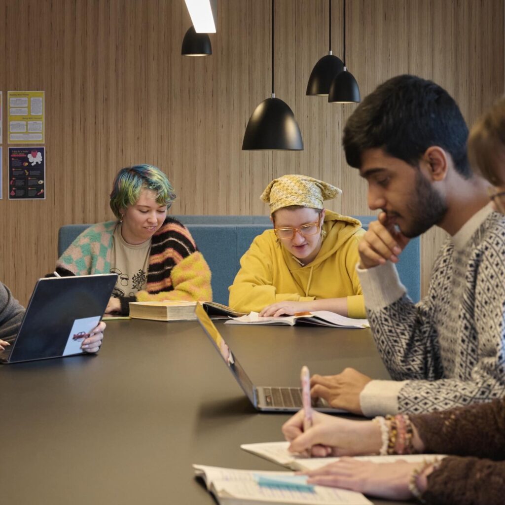 A group of five people sit at a table, studying and writing in notebooks. One person uses a laptop, another wears a yellow hoodie and headscarf, while another has colorful hair. Hanging lamps and a wood-paneled wall are in the background.