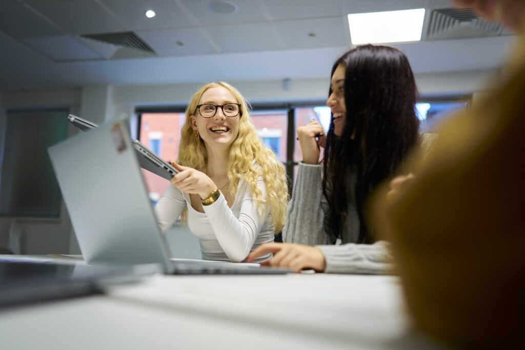 Two young women sit at a table with laptops, smiling and talking. One has long blonde hair and glasses, holding a tablet, while the other has long dark hair. They appear to be collaborating in a bright, modern room.