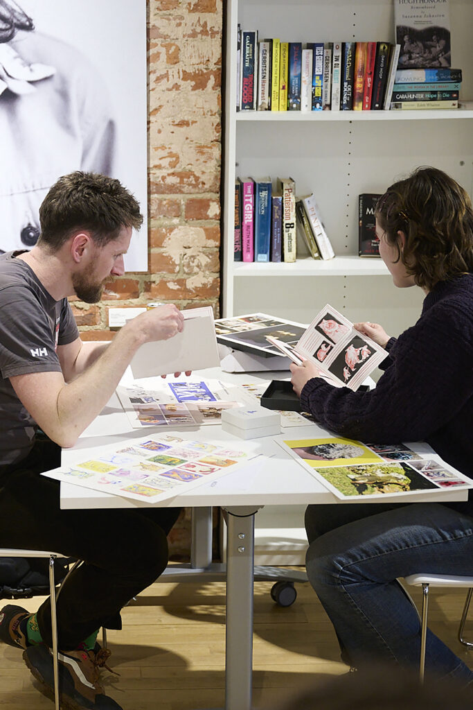 Two people sit at a table looking at printed photos and photo books, with colourful prints and books spread out. Behind them is a bookshelf filled with various books and a partial black-and-white portrait on the wall.