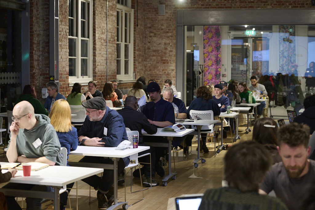 People sit in pairs and small groups at tables in a bright, modern room with brick walls, engaged in discussion or work. The atmosphere is lively and collaborative, with laptops and papers on tables.