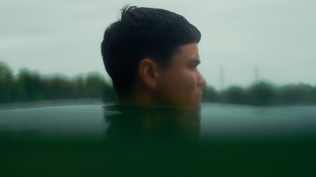 A young person with short dark hair is partially submerged in water, looking to the right. The background is blurred with trees and a cloudy sky visible. The mood is calm and contemplative.