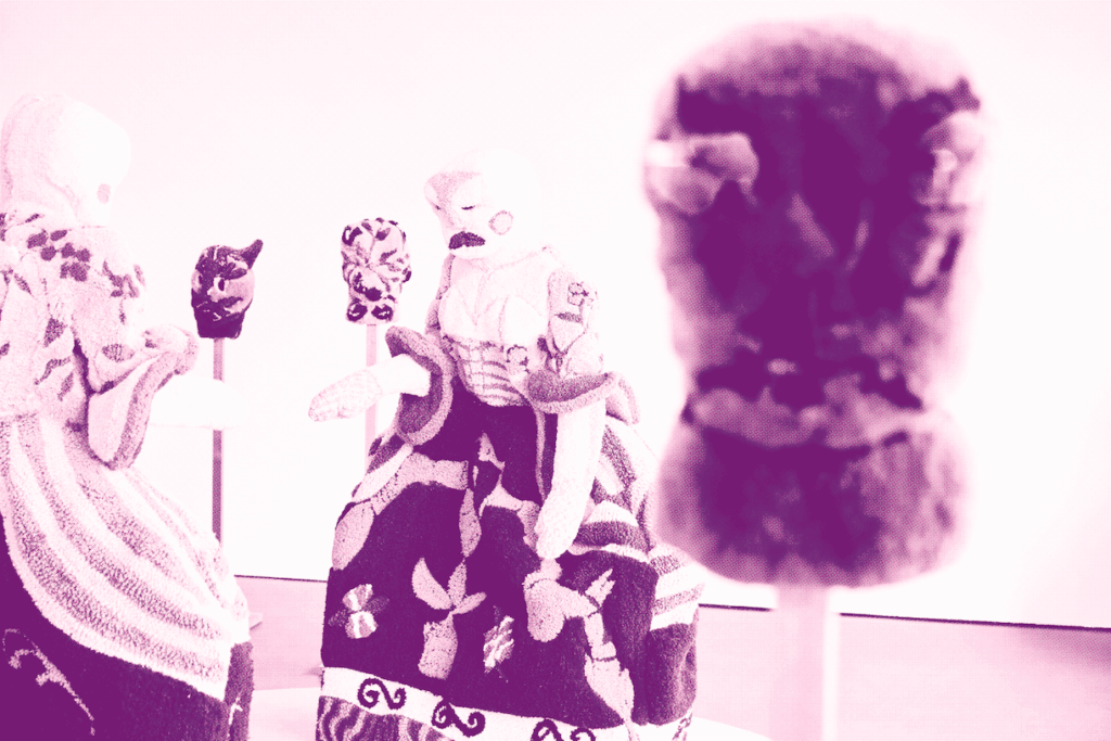 Three fabric puppets with detailed clothing and patterns are positioned in conversation, while a fourth puppet appears blurred in the foreground. The image is tinted with a soft pink hue.