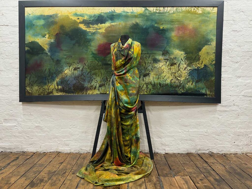 A mannequin draped in a green, yellow, and red patterned fabric stands in front of a matching painting, both set against a white brick wall and wooden floor.