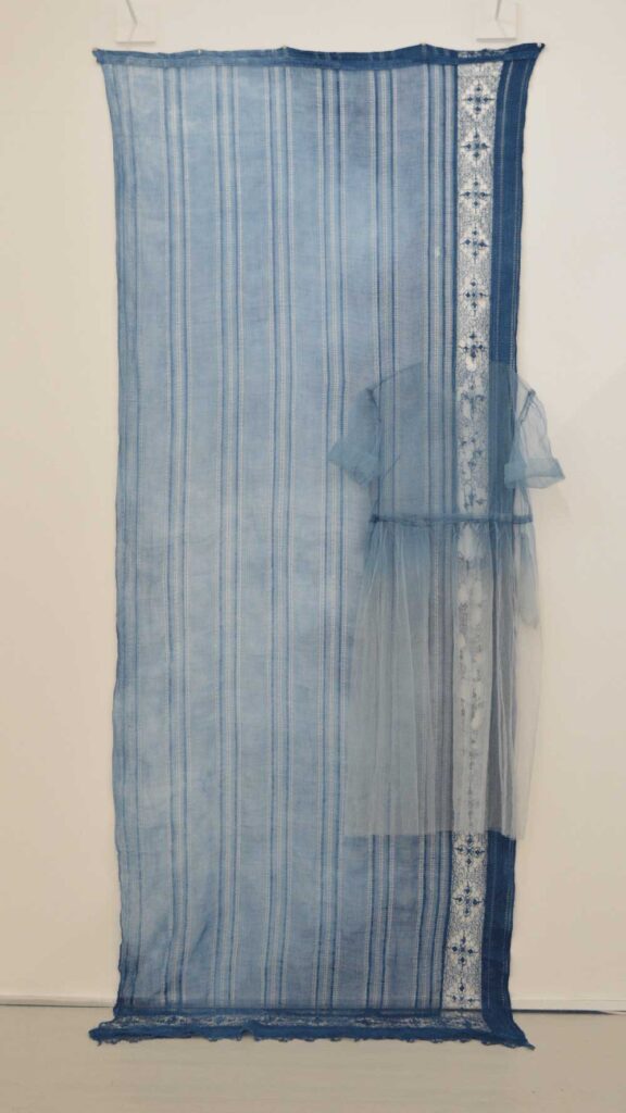 A sheer blue fabric with vertical stripes hangs on a wall, featuring an attached translucent blue dress and a decorative lace panel running down the right side.