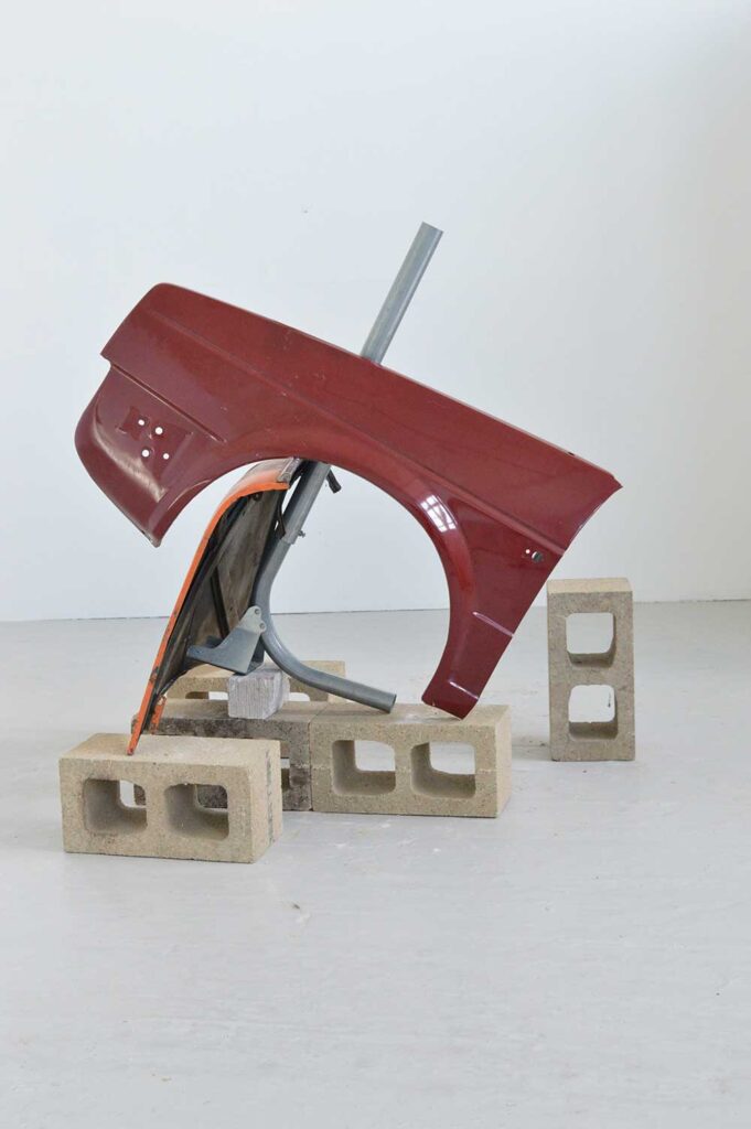 Parts of a car balance on concrete blocks, alongside metal rods, in a minimal, white-walled room, arranged as an abstract sculpture.