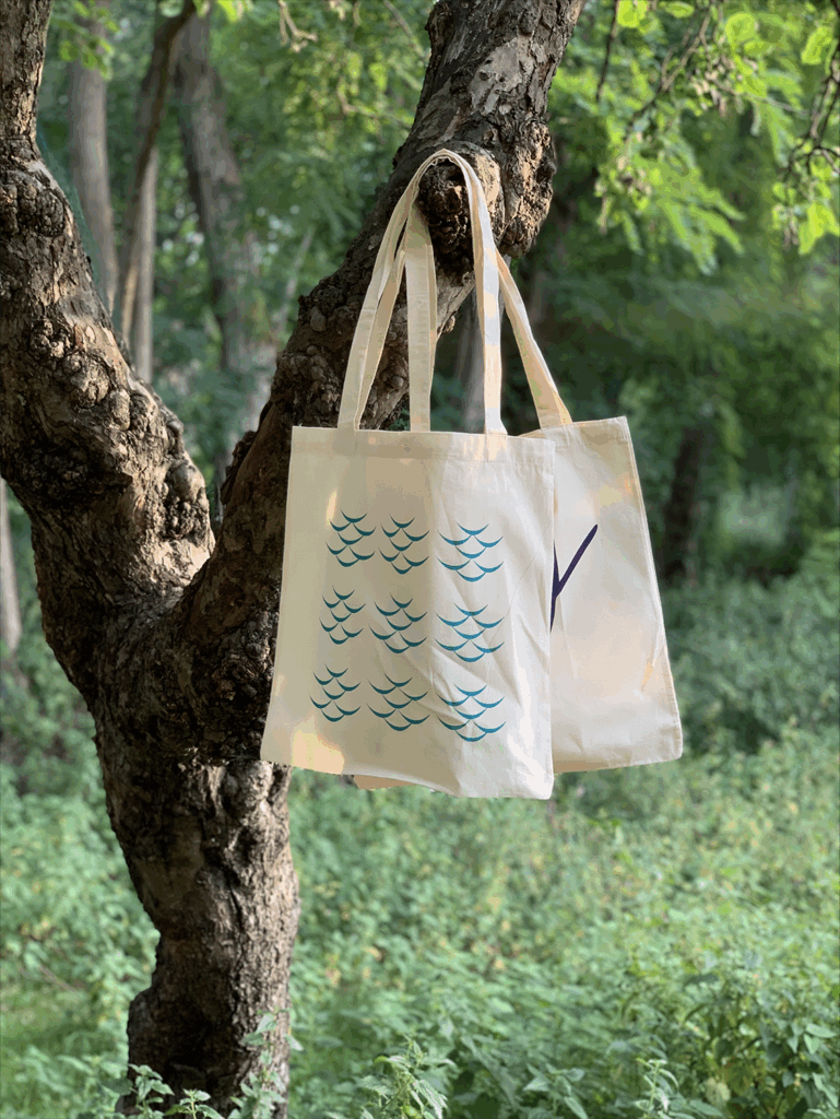 Two canvas tote bags hang from a tree branch. Sunlight filters through the leaves.
