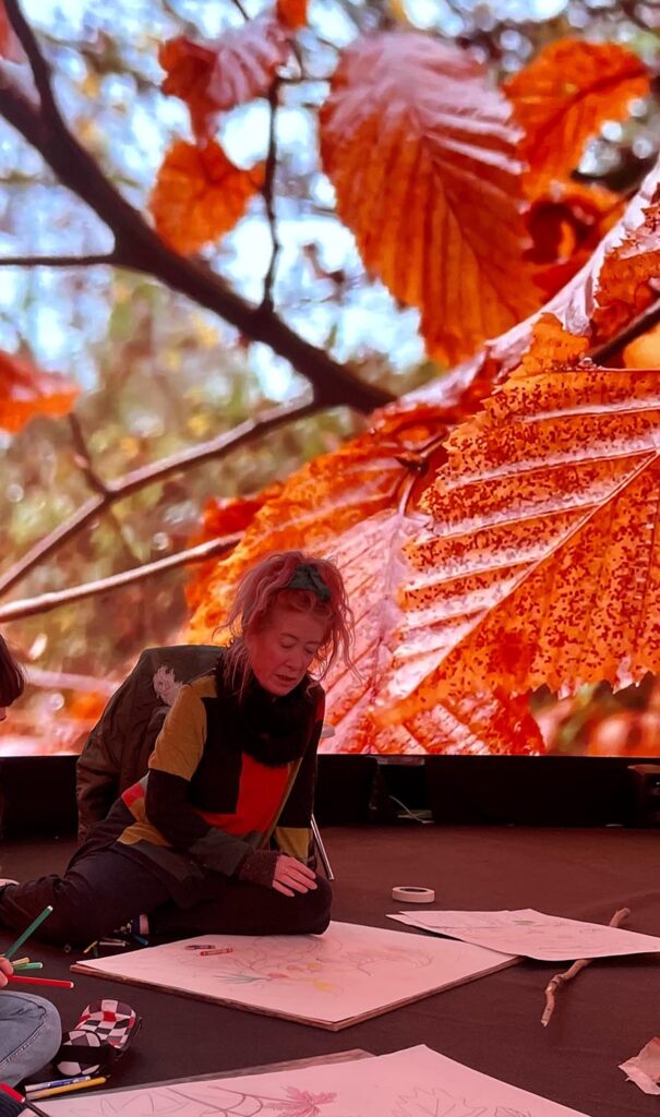 A person sits cross-legged on the floor, drawing on large sheets of paper. Behind them is a vivid, large screen displaying close-up autumn leaves in bright orange and red hues.