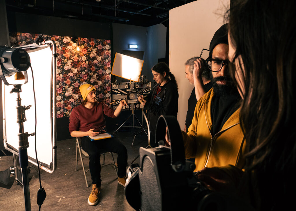 A film crew works on a set with bright lights, floral backdrop, and various equipment. One person sits on a chair holding papers. The scene has a creative, behind-the-scenes atmosphere.