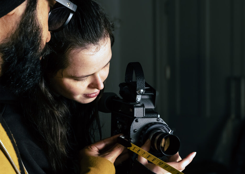 Two people closely examine a camera, with one person holding a tape measure against the camera lens and the other person looking intently at the measurement.