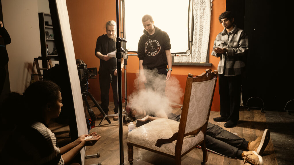 A film crew sets up a scene in a room with orange walls. One person lies on the floor exhaling smoke, while four others stand or crouch nearby, adjusting equipment and observing. A large chair is in the foreground.