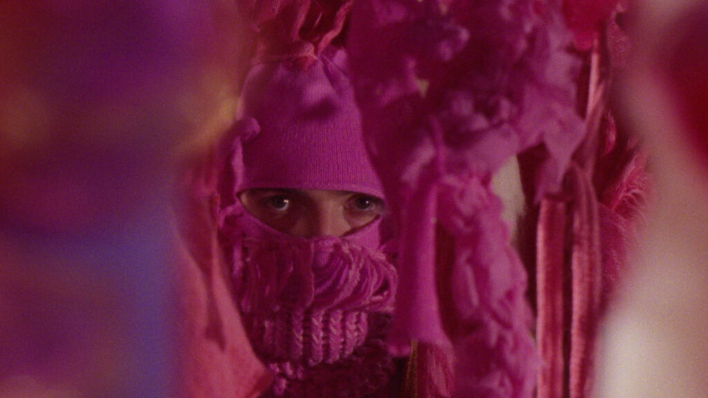 A person wearing a pink knit hat and scarf that covers most of their face peers through hanging, pink, textured fabric. Only their eyes are visible.