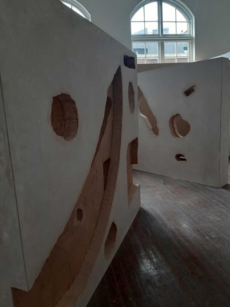Large sculptural structures with geometric cutouts stand in a room with wooden floors and tall arched windows, allowing natural light to highlight the textured surfaces of the installations.