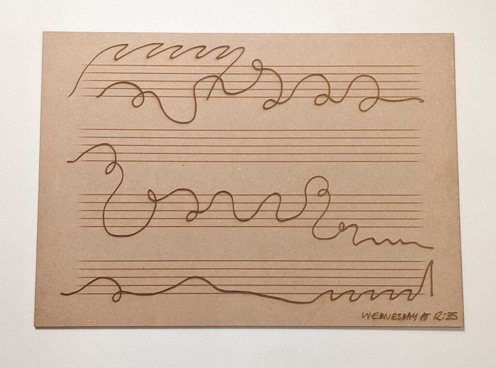 A piece of brown paper with four sets of parallel horizontal lines, resembling a musical staff, and wavy, irregular lines drawn across each staff in brown ink. The bottom right corner has text which reads: Wednesday at 12:35.