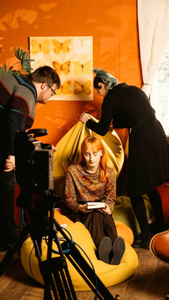A person with red hair sits on a yellow bean bag holding a book, while two people adjust the setting around them. A camera is set up in the foreground, and butterfly art decorates the orange wall behind them.