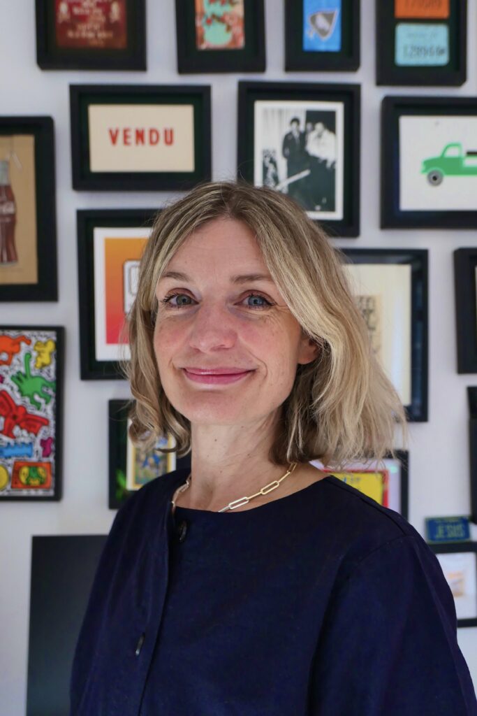 A woman with shoulder-length blonde hair and a navy blue top smiles in front of a gallery wall filled with colorful framed art and photographs.