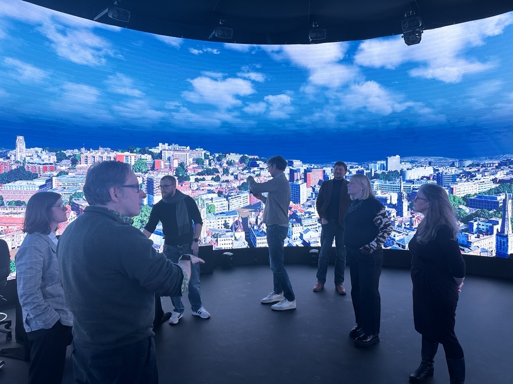 A group of people stands in front of a large, curved digital screen displaying a bright cityscape with blue skies and scattered clouds. Some are talking, while one person appears to be taking a photo.