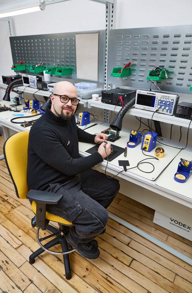 A man with glasses and a beard sits on a yellow chair at a workstation, holding a soldering tool. The desk has electronic equipment, soldering stations, and tools organized on shelves above.